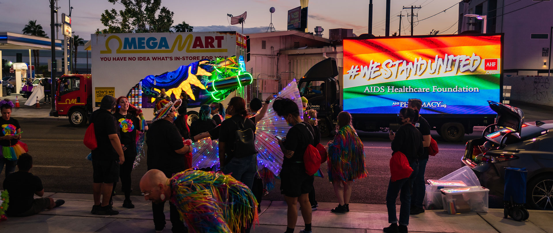 Omega Mart custom truck, with an open side, tubes, sun and cans lit up within, rolling in front of a digital billboard truck at the las vegas pride parade